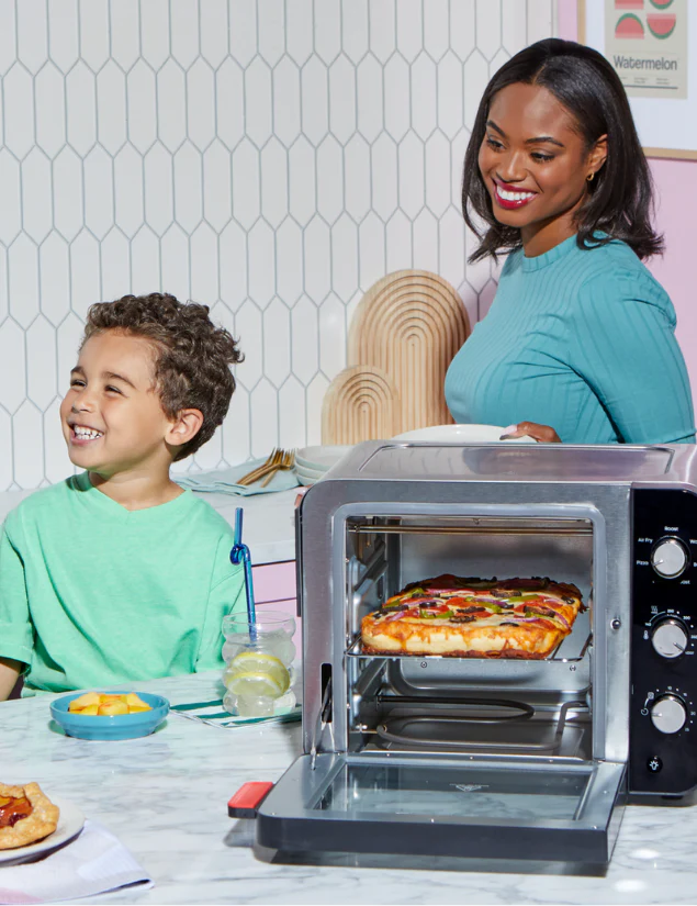 A woman in a teal top stands beside a young boy in a matching teal shirt, both smiling at the table. In front of them, a pizza bakes inside the Instant Pot® 4-Slice InstantHeat™ Mini Air Fryer Toaster Oven in Stainless Steel.